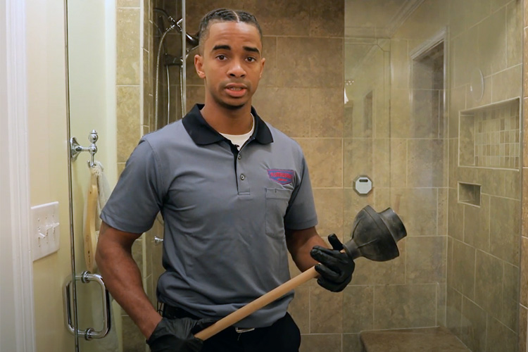 Experienced plumber given lesson on how to unclog toilet while holding plunger in a residential bathroom.